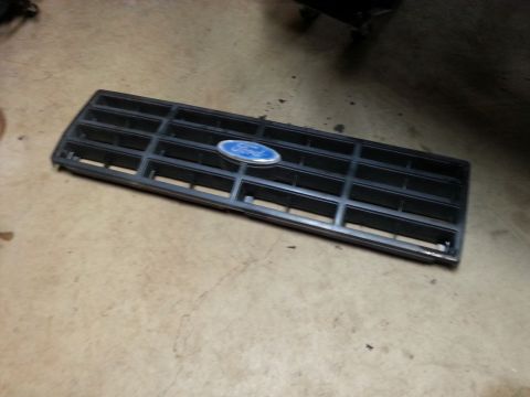 Removed black Ford truck grille with blue oval emblem lying on a garage floor