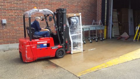 Operator driving a red Raymond forklift at a brick building loading dock