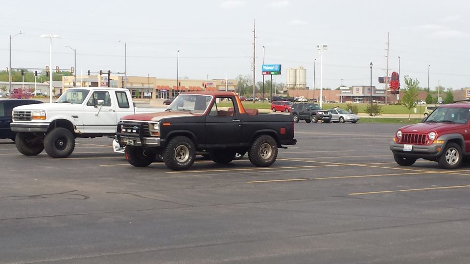 Black early Ford Bronco and lifted white F-series truck parked in a retail lot