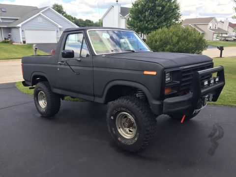 Matte black 1980s Ford Bronco with bull bar and aftermarket wheels parked on driveway.