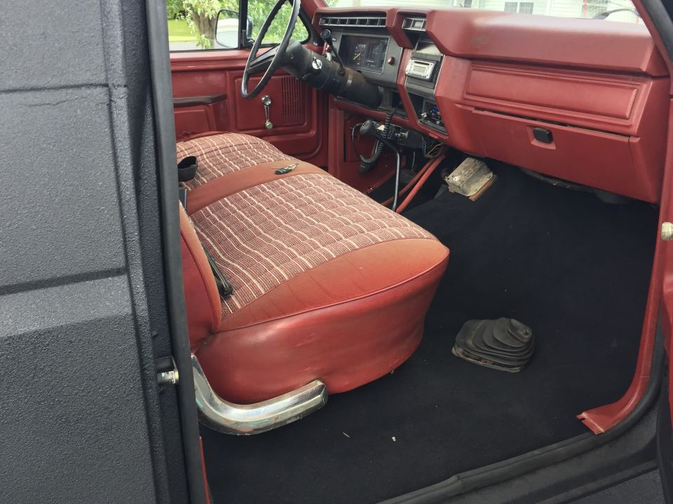 Interior of a vintage Ford Bronco showing red dashboard, plaid seats, and black carpet with shifter boot.