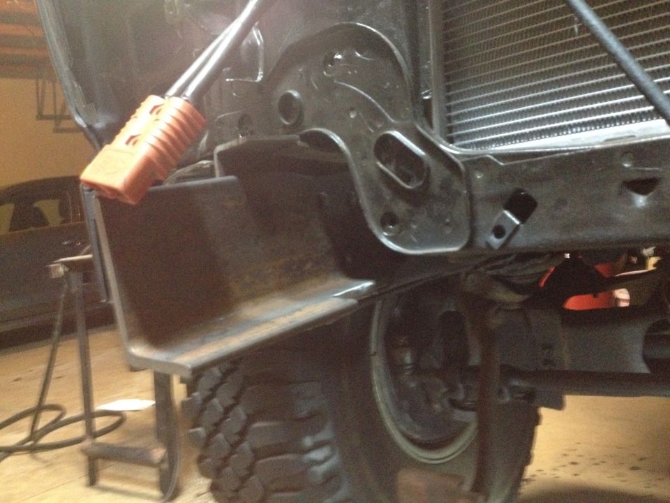 Underside of a vehicle showing frame, radiator, tire, and orange electrical connector.