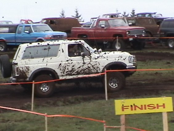 Mud-covered white Ford Bronco passing a yellow FINISH sign at a mud bog
