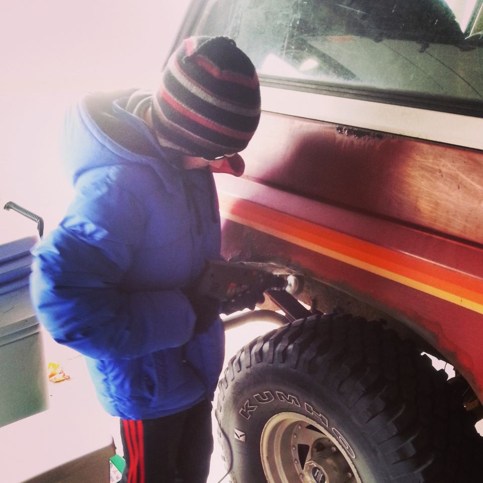 Person in blue jacket and knit hat working on the rear wheel area of a red and orange truck