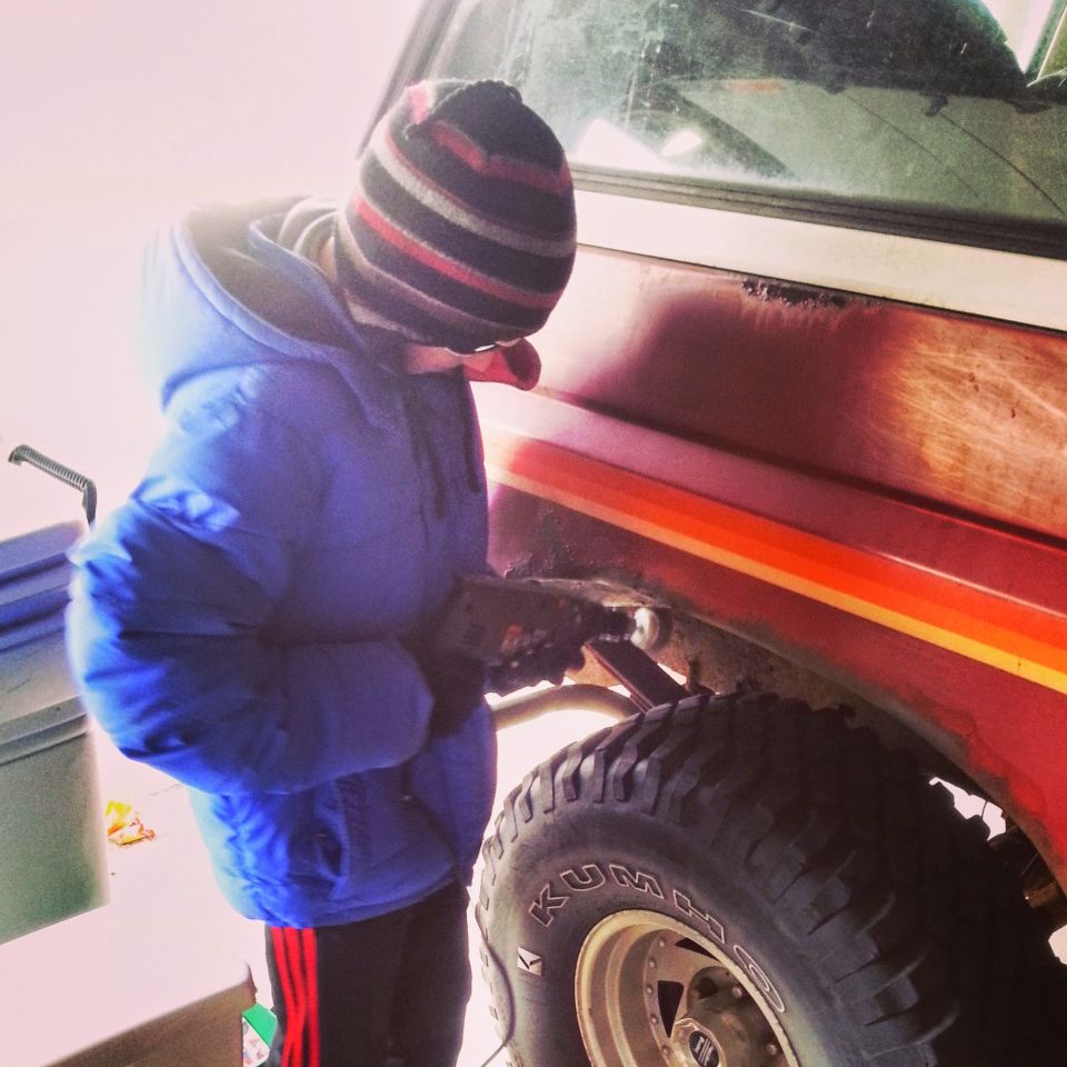 Person in blue jacket and knit hat working on the rear wheel area of a red and orange truck