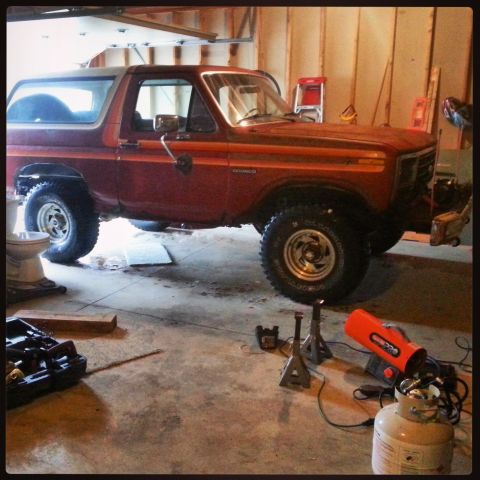 Red Ford Bronco on jack stands in a garage with tools and a propane heater