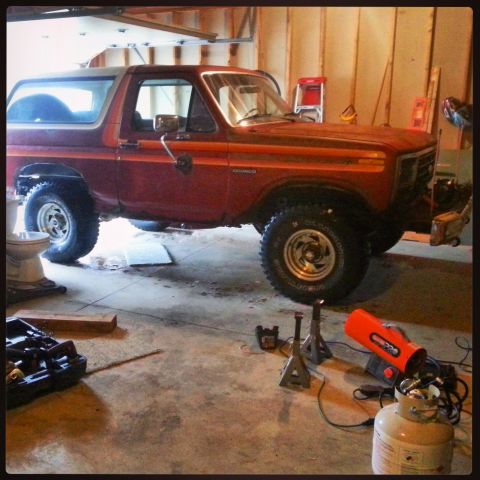 Red Ford Bronco on jack stands in a garage with tools and a propane heater