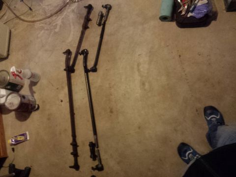 Two automotive steering linkage assemblies on a concrete floor with cans and sneakers.