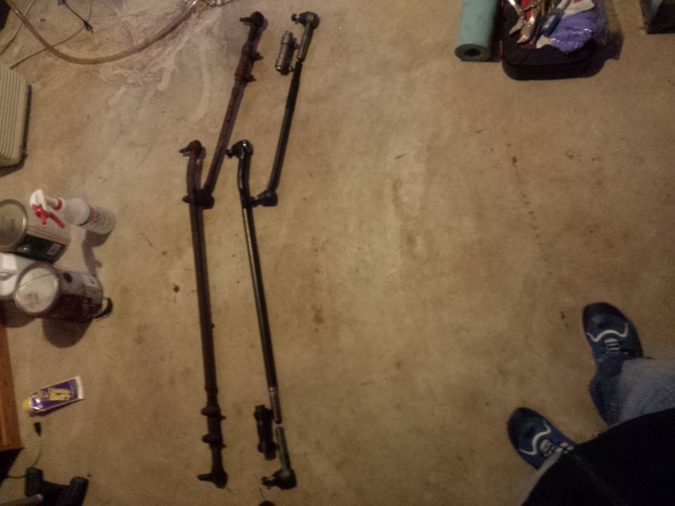 Two automotive steering linkage assemblies on a concrete floor with cans and sneakers.