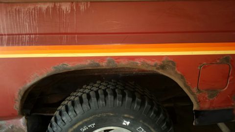 Close-up of rust damage on the rear wheel well of a red vehicle with racing stripes.
