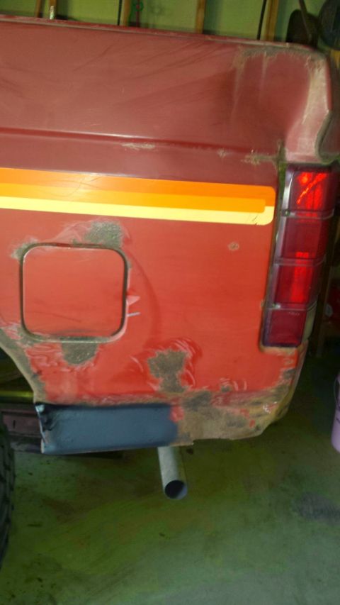 Rear quarter panel of a red vehicle with rust, peeling paint, fuel door, stripe, and taillight.