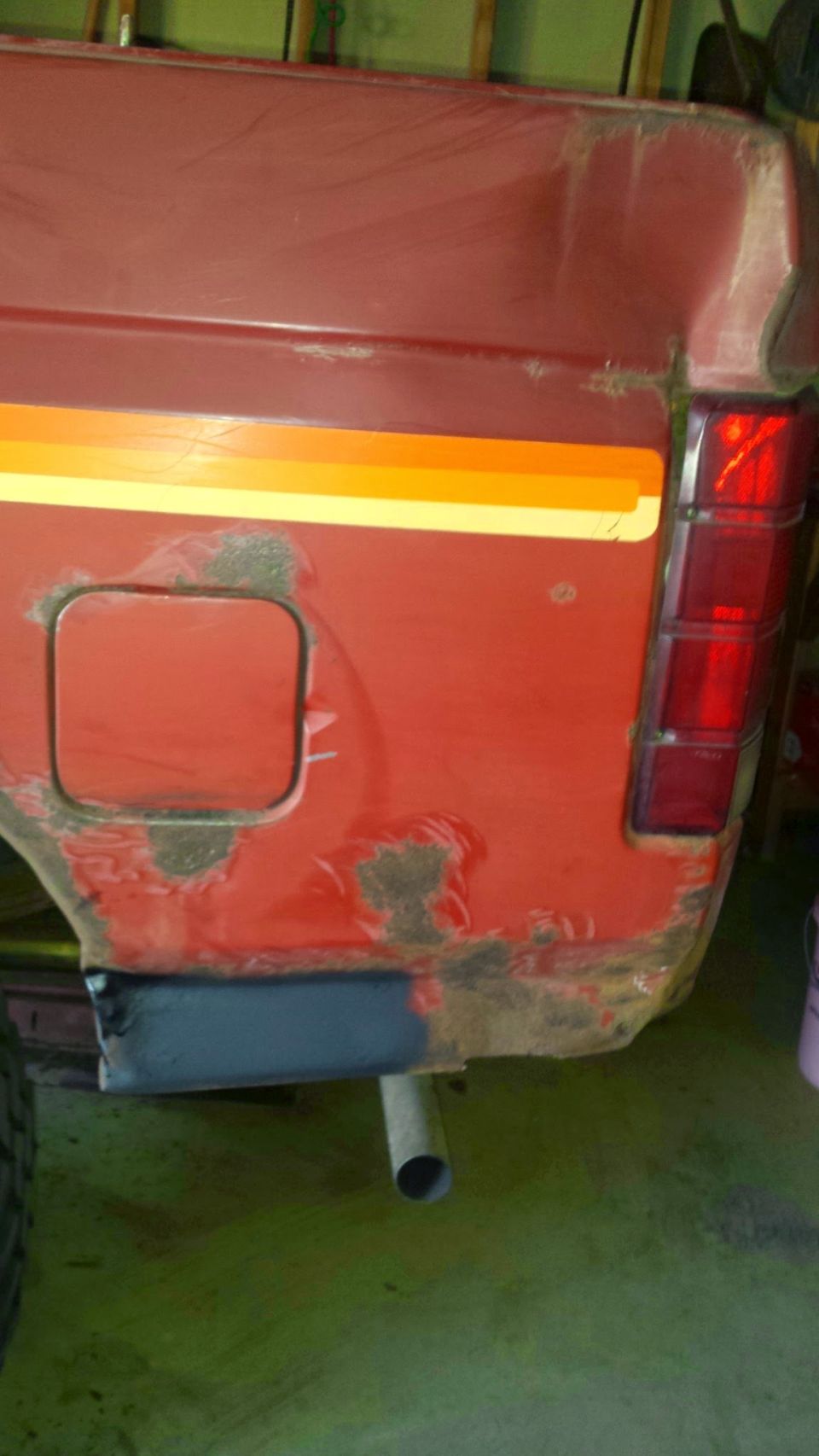 Rear quarter panel of a red vehicle with rust, peeling paint, fuel door, stripe, and taillight.