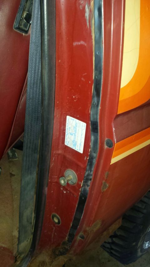 Close-up of red vehicle door jamb with seatbelt, sticker, rust, and exterior stripe decal.
