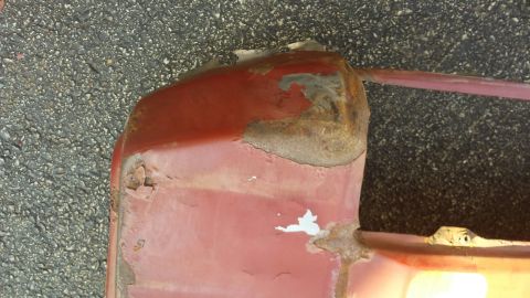 Rusted red vehicle panel corner with peeling paint on asphalt.