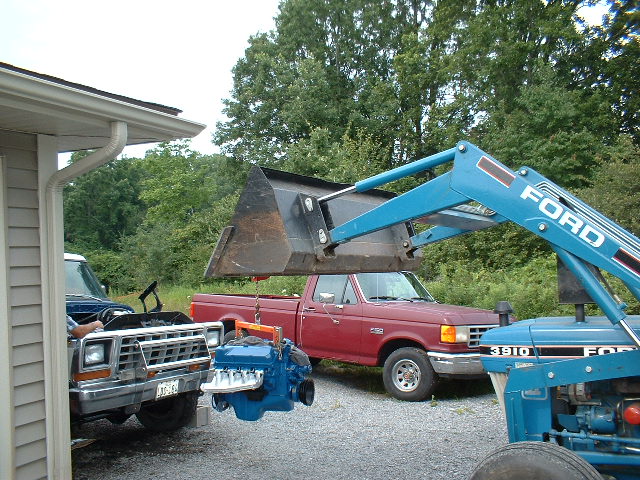 Red Ford pickup truck with engine being lowered by loader; front of another Ford truck visible