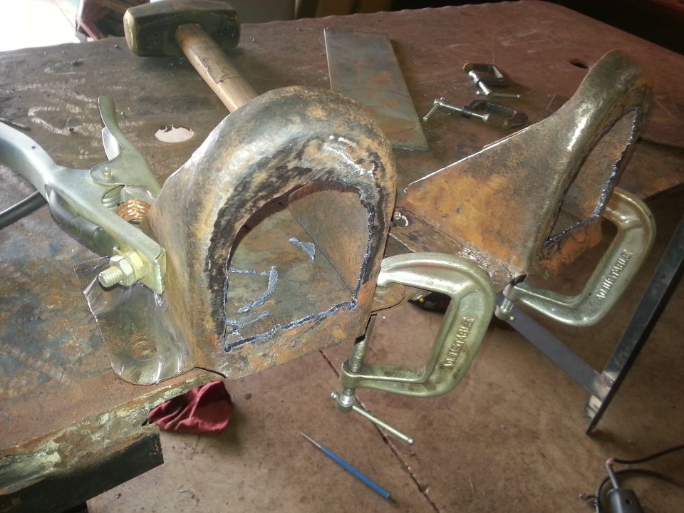 Two rusty metal parts clamped to a workbench, one U-shaped and one wedge-shaped, with cut lines and welding marks.