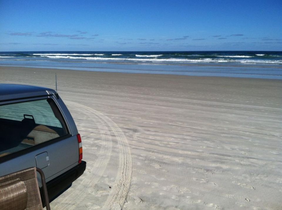 Silver station wagon parked on a sandy beach with ocean waves in the background. Tire tracks are visible in the sand