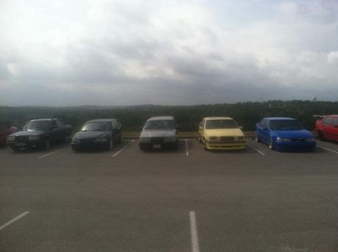 1991 Volvo 940 - A row of cars are parked in an outdoor lot with a wooded area in the background.