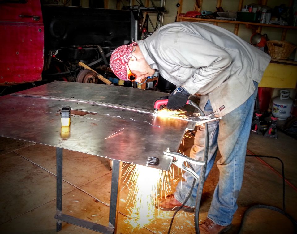 Man welding metal sheets in a garage with a red vehicle in the background