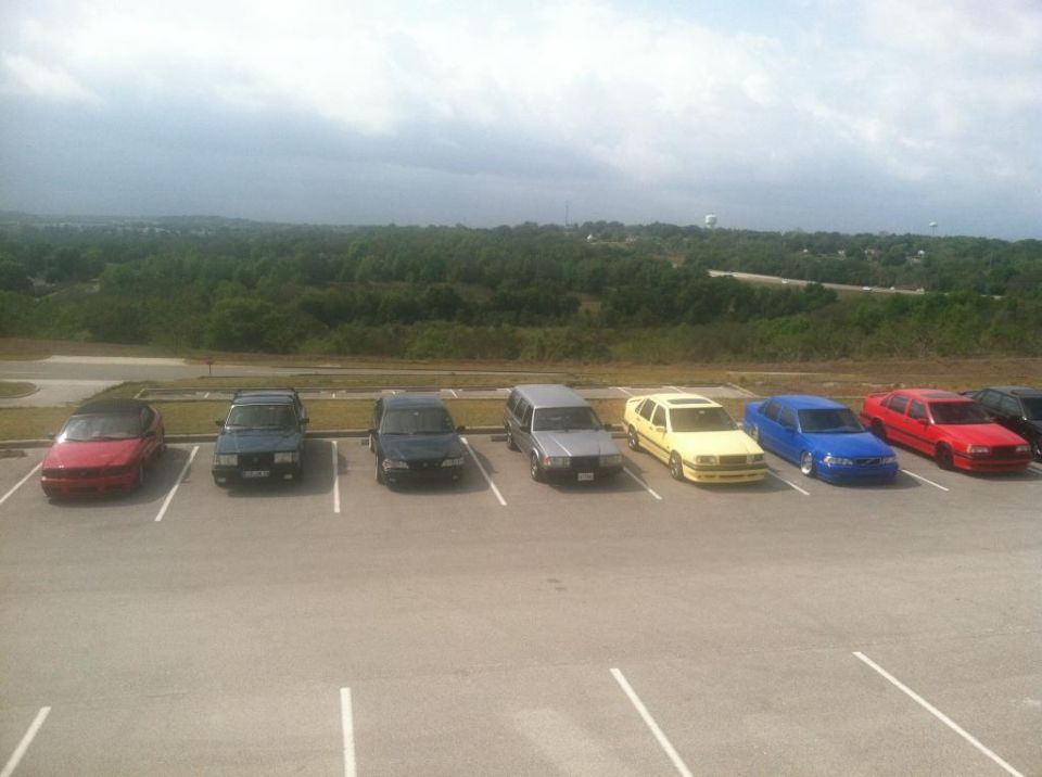Several cars parked in a lot with a wooded area and cloudy sky in the background