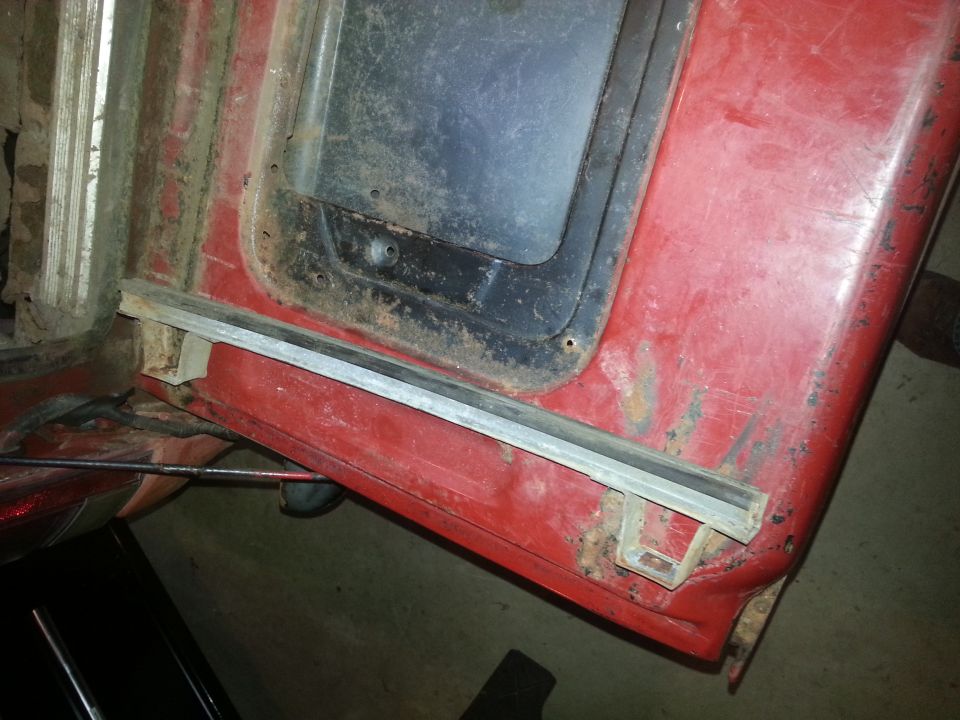 Red vehicle door frame with a black window opening and visible rust, showing a metal trim piece