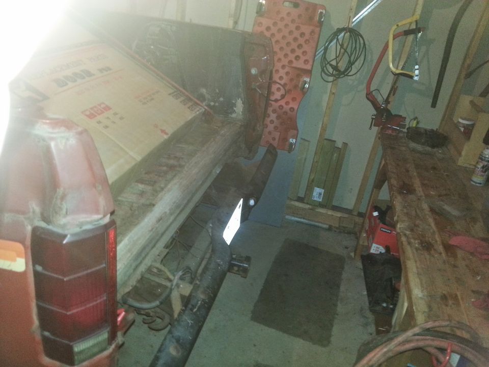 Rear of a red SUV, possibly a Ford Bronco, with a large cardboard box in the cargo area. Tools and a workbench are visible in the background