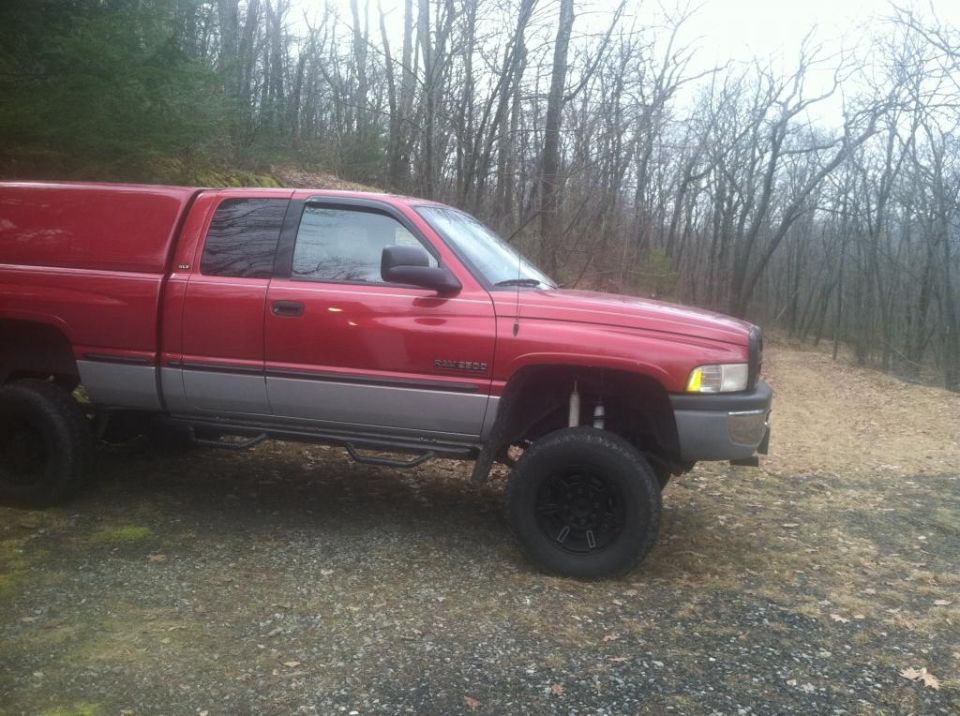 Lifted red and silver 1999 Dodge Ram 2500 pickup truck with camper shell and black wheels, parked outdoors.