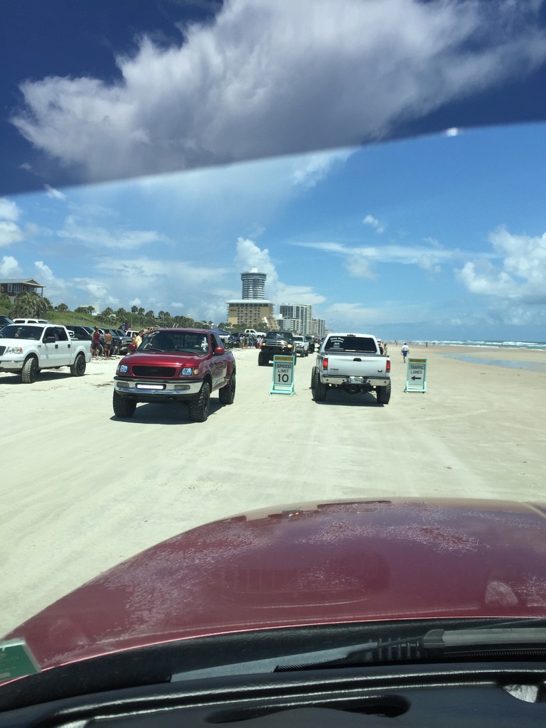 Pickup trucks on a beach, with a red truck in the foreground and others in the background.