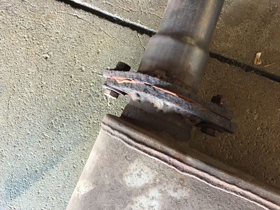 Rusted exhaust pipe connection with copper gasket and bolts on a concrete surface.