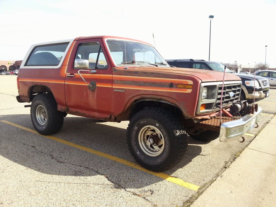 Rusty red 1980s Ford Bronco with white top and racing stripes, parked in a lot.