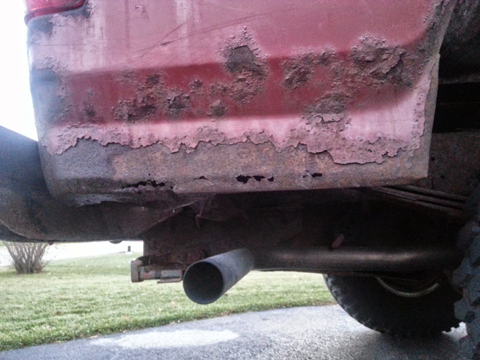 Close-up of heavily rusted red vehicle quarter panel with exhaust pipe.