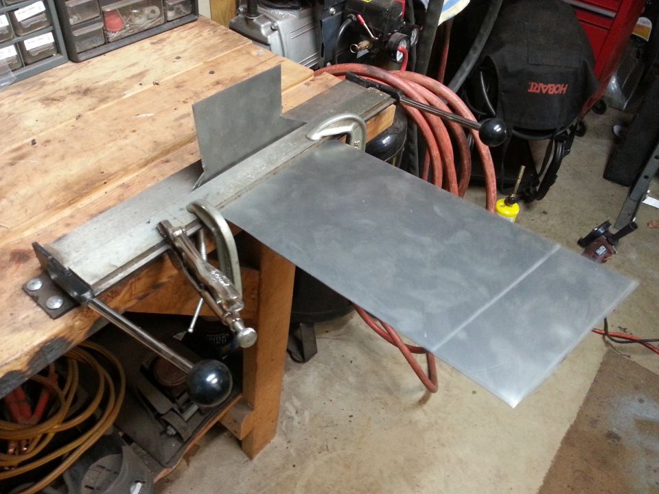 Metal sheet being bent on a workbench with clamps and a jig.