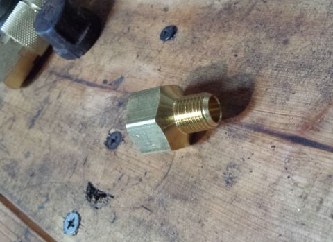 Brass fitting with hexagonal ends and threaded connectors on a wooden workbench.