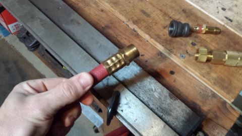Hand holding a red hose with a brass fitting. Other fittings on a wooden surface.