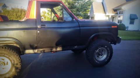 Driver's side view of a dark Ford Bronco with off-road tires and a striped roof.