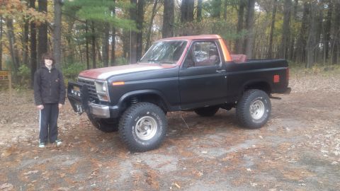 Lifted black and red Ford pickup truck with orange accents, parked in a wooded area.