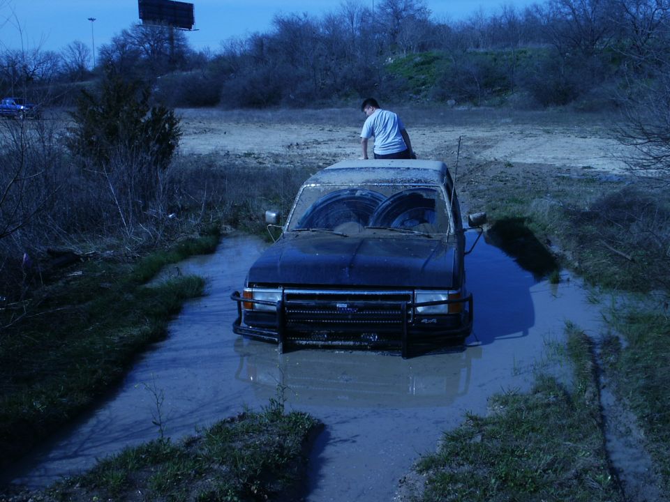 Muddy Ford Bronco with grille guard stuck deep in a mud hole, person sitting on the roof