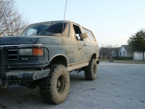 1991 Ford Bronco - Low-angle 3/4 front view of a lifted dark blue Ford Bronco caked in dried tan mud from bumper to roof, parked on a suburban street. It wear…