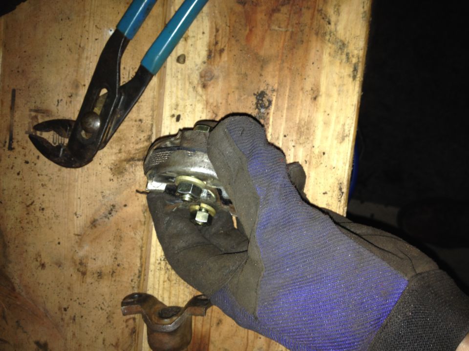 Gloved hand holding a metal part with nuts and bolts, pliers above. Wood background