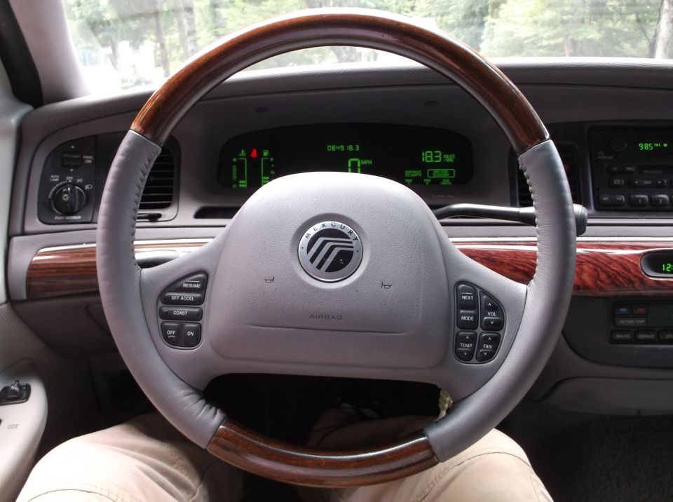 Interior view of a car steering wheel, dashboard, and center console with wood trim and digital gauges.