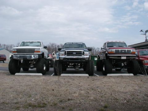 1985 Ford Bronco - Three lifted pickup trucks, including a red Ford F-Series, are parked in a lot under a cloudy sky.