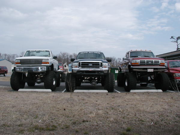 Three lifted pickup trucks parked side-by-side, front view. Red truck in foreground