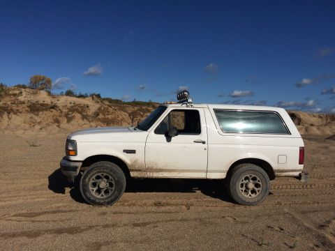 1993 Ford Bronco - A white Ford Bronco is parked on a sandy terrain under a bright blue sky with scattered clouds. The vehicle is viewed from the side, showin…