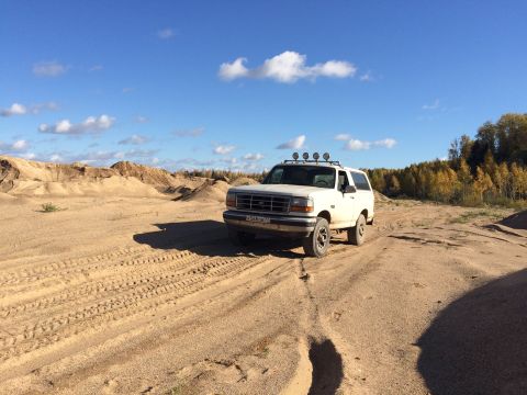 1993 Ford Bronco - A white 1993 Ford Bronco is parked on a sandy, off-road terrain under a blue sky with scattered clouds. The vehicle has four auxiliary ligh…