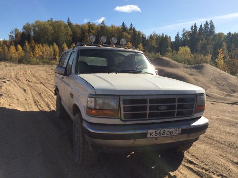 1993 Ford Bronco - A white 1990s Ford Bronco is parked on a sandy terrain with tire tracks visible. The vehicle is facing forward with a forest and a clear bl…