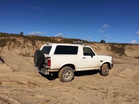 1993 Ford Bronco - A white 1993 Ford Bronco is parked on a sandy terrain with a steep, layered dirt embankment in the background under a clear blue sky.
