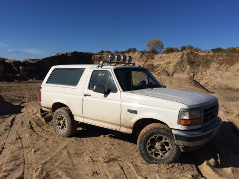 1993 Ford Bronco - A white 1993 Ford Bronco is parked on a sandy terrain with tire tracks visible in the foreground. The vehicle has four auxiliary lights mou…
