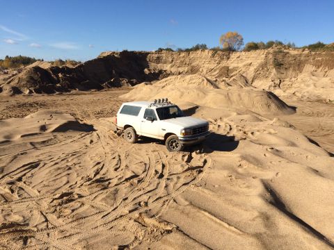 1993 Ford Bronco - A white 1993 Ford Bronco is parked on a sandy dune in a quarry under a clear blue sky. The vehicle has a roof rack with multiple lights mou…