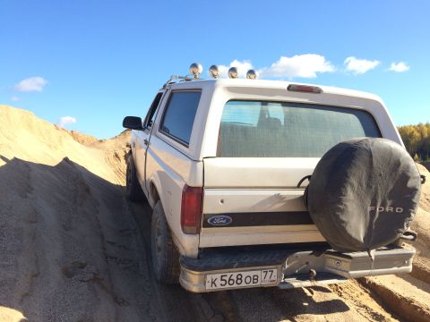 1993 Ford Bronco - Rear 3/4 view of a white Ford Bronco driving on a sandy hill under a blue sky with scattered clouds. The vehicle has a spare tire cover on…