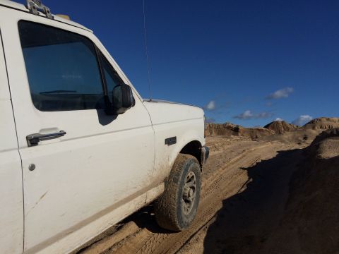 1993 Ford Bronco - A white Ford Bronco is parked on a dirt road with sandy hills and a bright blue sky in the background. The side of the vehicle and its fron…